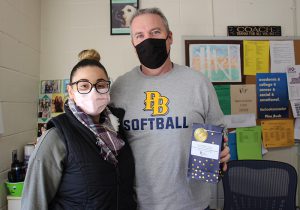 A man in a PB Softball tshirt stands holding a black bag with stars on it. He has short hair and is wearing a black mask. He is standing next to a woman with a bun on top of her head, wearing a black vest, plaid scarf and a pink facemask and glasses.