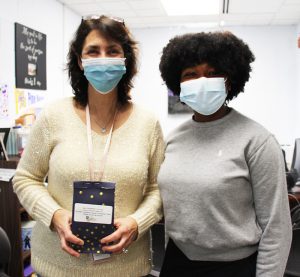 A woman with shoulder-length dark hair is wearing a cream colored sweater and a blue mask. She is holding a black bag with stars on it. She is standing next to a high school student in a gray sweater, wearing a blue mask. Se has shorter curly hair.