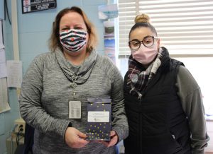 A woman in a gray sweater wearing a blue and white striped face mask, with chin-length blond hair holds a black bag with stars on it. She is standing next to a woman with a bun on top of her head, wearing a black vest, plaid scarf and a pink facemask and glasses.