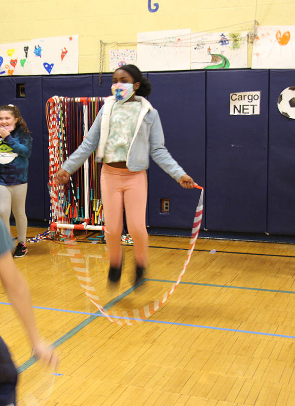 An elementary age girl, wearing pink pants and a light blue jacket and mask jumps rope in a gym.