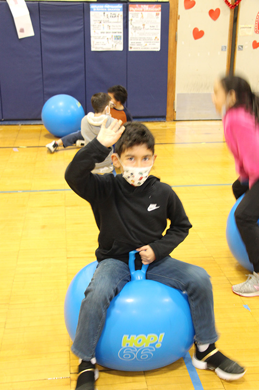 A boy with short dark hair sits on a blue bouncy ball with a handle. He is wearing a dark sweatshirt and light colored mask.