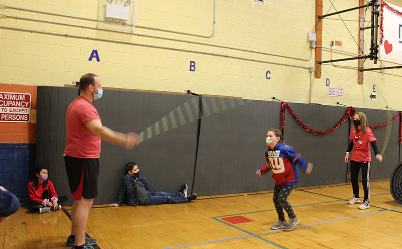 An elementary age girl, wearing a mask and a red and blue NY Giants shirt jumps rope while two adults, a man on the left and a woman on the right, turn the rope for her. They are in a gym,