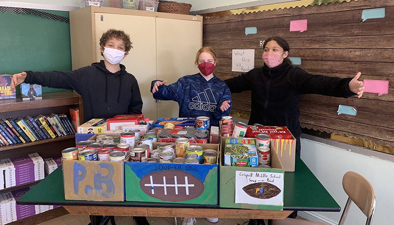 Three middle school students standing with their arms out. In front of them are boxes of canned goods and other non-perishable good items in boxes with PB on them, a football that says soup-er bowl and Crispell Middle School Soup-er bowl