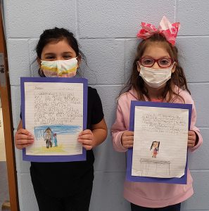 Two small girls wearing white masks stand against a wall holding up a piece of paper. On the paper is a picture they drew and a story they wrote.