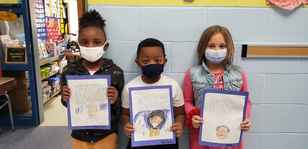 Three second-grade students wearing face masks, stand against a wall holding up a piece of paper. On the paper is a picture they drew and a story they wrote.