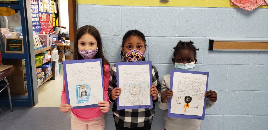 Three girls wearing face masks, stand against a wall holding up a piece of paper. On the paper is a picture they drew and a story they wrote.