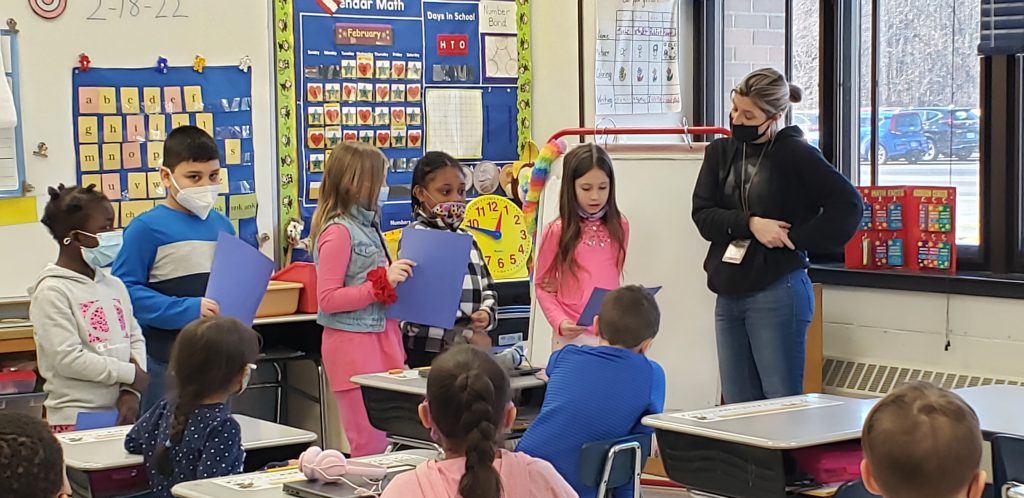 In a classroom a woman stands at the front of the class looking at a line of second-grade students. There five children along the side of the room and several more sitting at their desks. The children standing have papers in their hands.