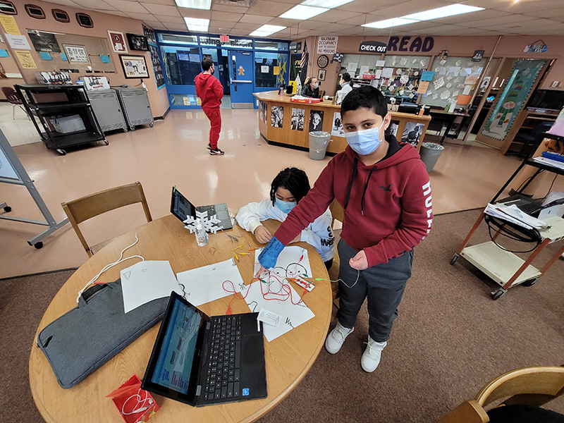 A students wearing a red hoodie stands at a desk in the library. There are people and books and shelves in the background. Another person is sitting at the table nearby. The student has a wire coming from the tablet and drawings on the table.