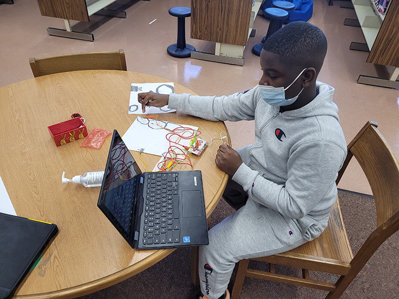 A student with very short brown hair sits at a table in the library. A tablet is open on the table and an orange wire is attached to it. He is holding the wire in one hand and has his finger from the other hand on a drawing on a piece of paper.