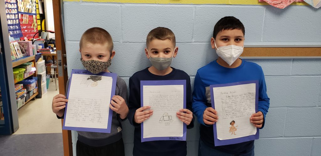 Three boys with short hair, wearing face masks, stand against a wall holding up a piece of paper. On the paper is a picture they drew and a story they wrote.