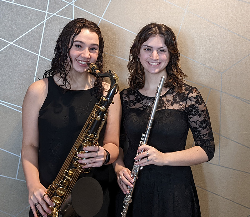 Two young women, both with long dark hair, wearing black dresses hold musical instruments - a sax and flute.