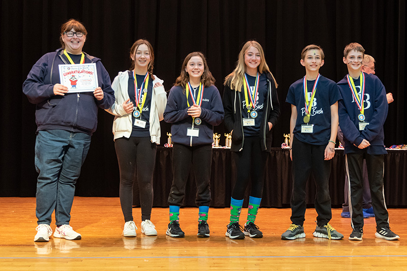 Five children and one adult stand on a stage. They all have medals around their necks and blue shirts with PB Pine Bush on them. They are all smiling.