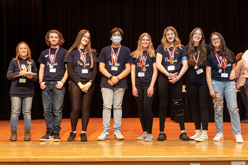 A woman is holding a trophy on the right. There are seven older students to her left. They are wearing medals around their necks. They all have navy blue tshirts with PB Pine Bush on them.