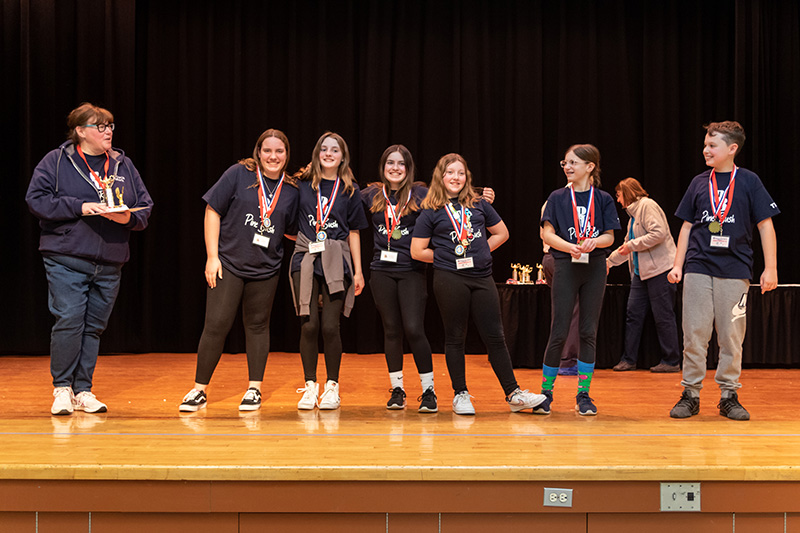 Six students and an adult stand on the stage. They are all wearing navy blue tshirts that say PB Pine Bush on them. They all have medals around their necks.