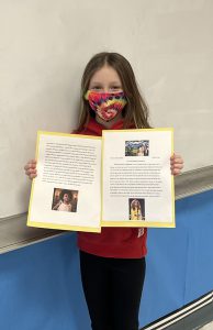 A fifth grade girl wearing a tie dye mask, long brown hair, holds up her essay.