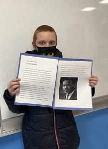 A fifth grade boy with very short light hair holds up his essay of Martin Luther King Jr. It has a picture of  King on it.