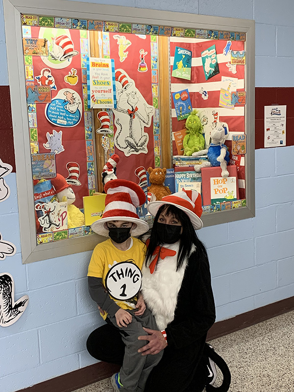 A large glass case on the wall filled with Dr. Seuss books and photos. In front, squating down, is a little boy with a red and white top hate on and a shirt that says Thing 1. Next to him is a woman dressed as the cat in the hat. Both have black masks on.