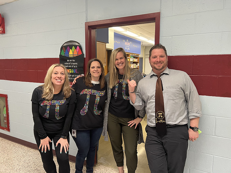 A man on the right and three women next to him all are wearing te pi symbol. The women have navy blue shirts with the pi symbol in different colors while the man has a pi symbol on his tie with the word inspire.