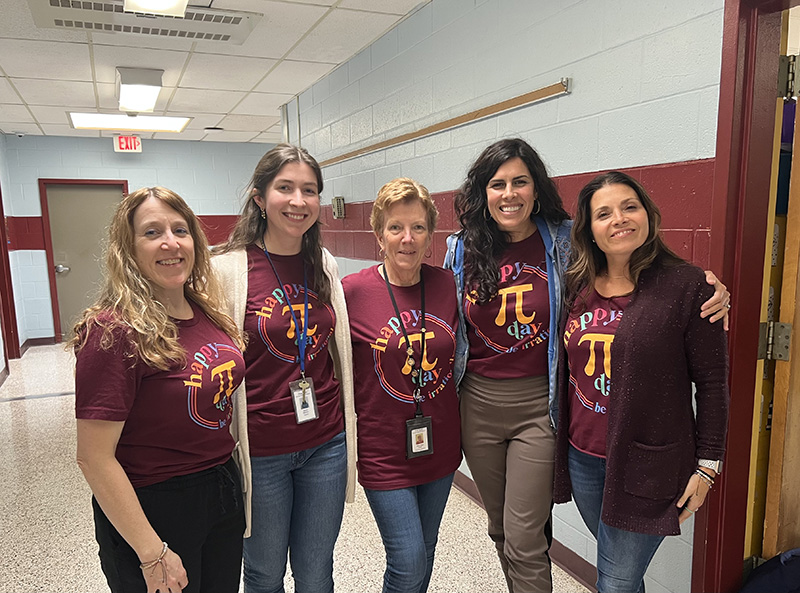 Five women stand together. All are wearing maroon shirts with the pi symbol in the center in gold.