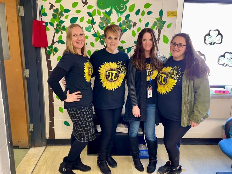 Four women stand together. All are wearing navy blue shirts with yellow sunflowersin the center of them. In the center of the sunflower is the pi symbol.