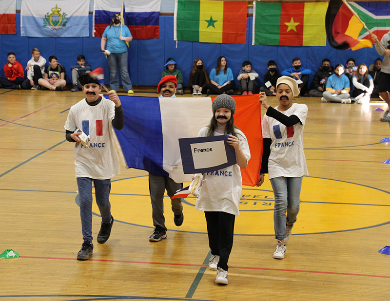 Four middle school students in a gymnasium carrying the flag of France. They have it on their shirts too. And some are dressed as Frenchmen, with berets and mustaches.