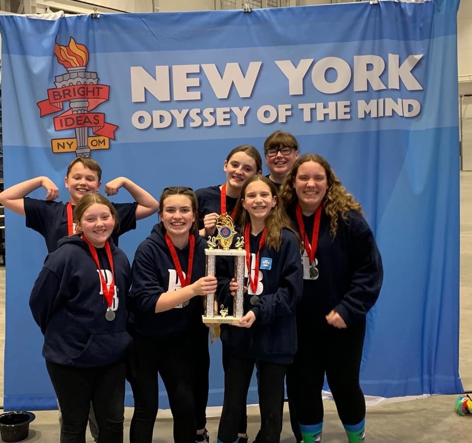 Six middle school students and a woman all smiling and wearing navy blue sweatshirts with the letters PB in white on them. They have red ribbons around their necks. Two of the students are holding a large trophy. One student on the left is holding up his arms as if to show his miscles.