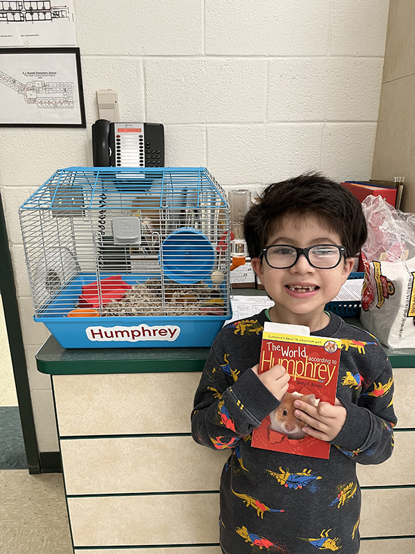 A little boy with black hair and glasses is smiling holding a red book. Next to him is a hamster cage with a hamster in it.