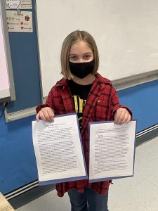 A fifth grade student with chin length blond hair, wearing a black mask and red and black plaid shirt, holds up an essay.