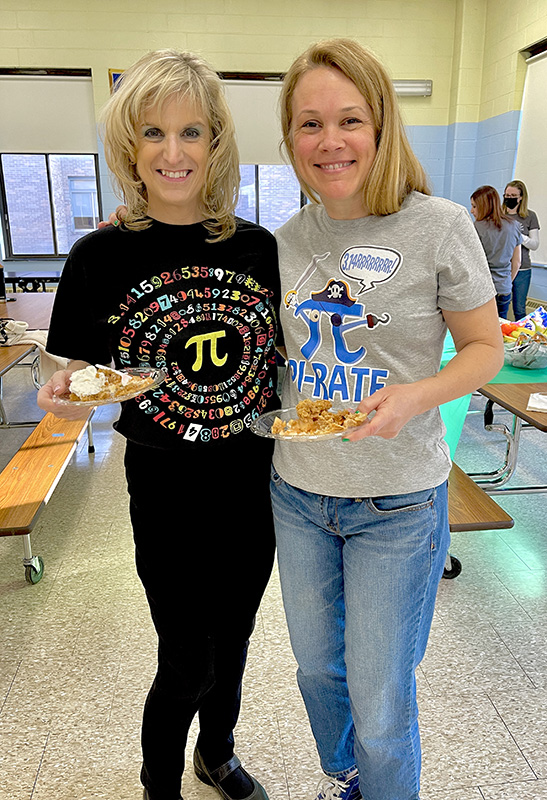 Two women stand together holding plates with pie on them. They are also wearing shirts with the pi symbol on them.