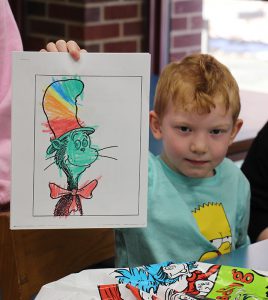 A kindergarten boy with red hair, wearing a light green shirt holds up a picture he colored of the Cat in the Hat. 