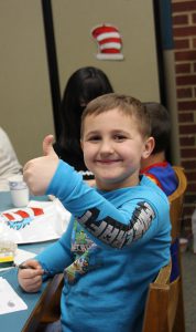 A kindergarten boy wearing a long sleeve blue shirt smiles and gives thumbs up.