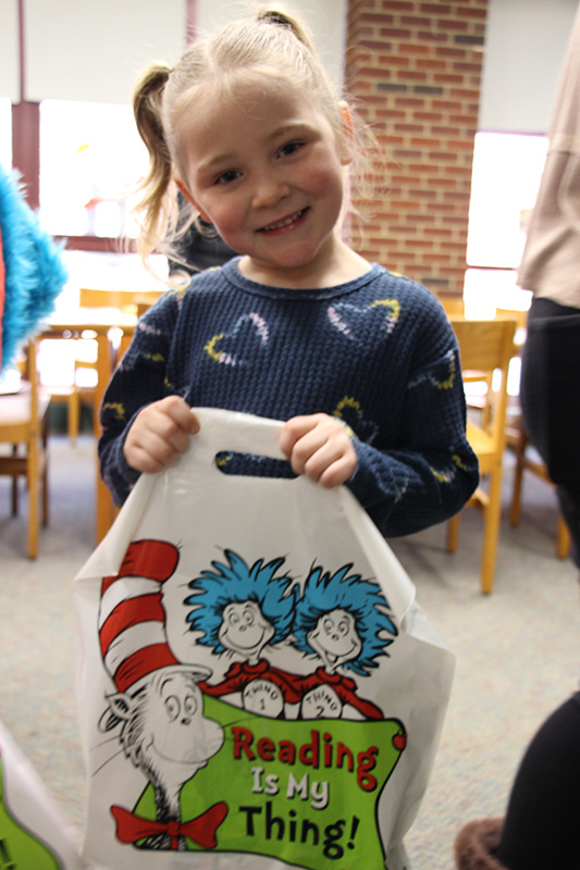 A kindergarten girl with blonde hair in ponytails, wearing a blue shirt with hearts on them holds a plastic bag with a picture of the Cat in the Hat on it and Thing 1 and Thing 2. She is smiling.