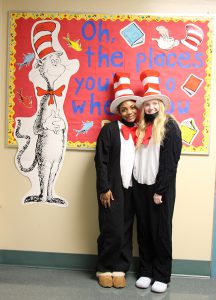 Two high school students dressed in black and white cat costumes with tall red and white hats on stand in front of a bulletin board that says Oh the places you'll go when you read, with a picture of the Cat in the Hat on it.