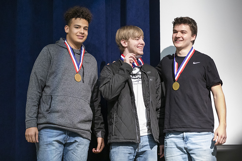 Three male high school students stand on a stage with a blue curtain behind them. They each have a medal around their neck with a red white and blue ribbon. They are all smiling.