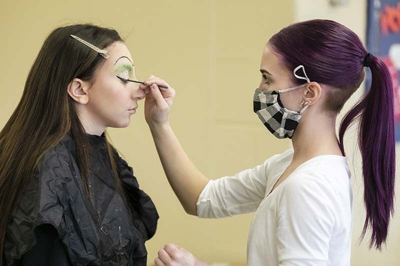 Two young women stand facing each other. The young woman on the right is wearing a black and white checked face mask. She is