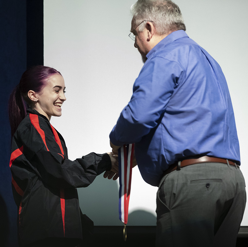 A young woman dressed in a black and red blouse smiles as she shakes hands with a man in a blue shirt and gray pants. He is holding a medal on a red, white and blue ribbon to give to her.