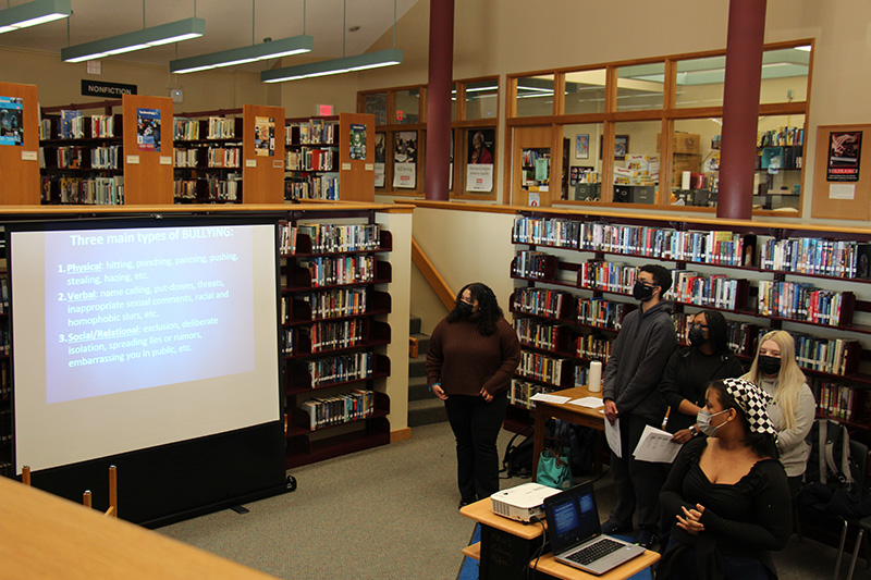 The setting is a library. There is a screen at the front of the room with a list of  three types of bullying - Physical, verbal and social/relational. There are a handful of high school students on the side looking at the screen.