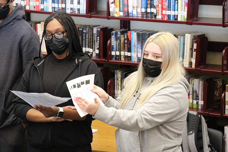 Two high school students leading a forum. One on the right, with long blonde hair, holds a piece of paper she is showing to a group. The young woman on the left, who has long dark braids, is reading from a paper.