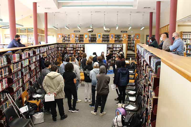 A large group of ninth-grade students is in a section of a library, all looking toward a screen at the front of the room.