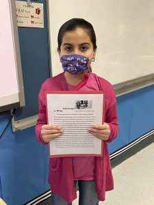 A fifth grade girl wearing a printed blue mask, long brown hair pulled back in a ponytail, pink shirt, holds up her essay.