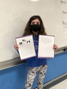 A fifth grade girl wearing a black mask, blue shirt and printed pants, long brown hair, holds up her essay.