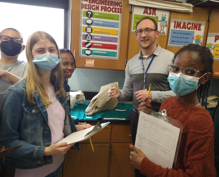 a man with glasses, wearing a gray shirt stands with four middle school students with skulls in the background. The students are holding clipboards.