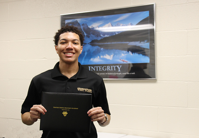 A young man, high school senior, smiling broadly. He s wearing a black polo shirt that says West Point on it. Behind him is a poster with the word Integrity on the bottom. He is holding a black folder with gold imprint. It says United States Military Academy West Point , N.Y.