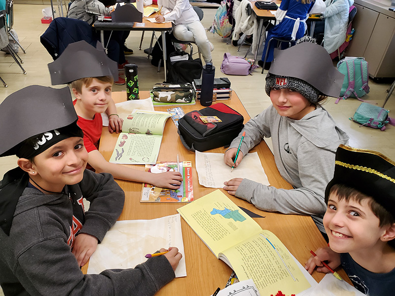 Four boys sit at a table writing on  colonial paper. They are all wearing colonial hats.