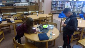 Two middle school students are at a round table in a library. One is leaning on the table and the other is standing working with something in his hands. There is a laptop on the table, a water bottle and other pieces of robotics materials.