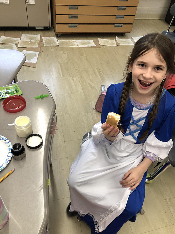 A girl with long dark hair holds a piece of bread with homemade butter on it. She is wearing a blue and white colonial costume.