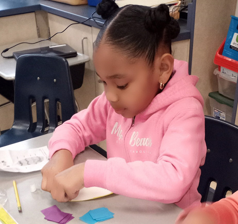 A second-grade girl, whose dark hair is pulled up into two twists on the sides of her head, sits at a desk. She is making something with blue and purple paper. She is wearing a pink sweatshirt.