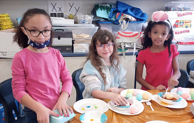 Three second-grade girls color eggs. They are all smiling. Girl on the left is wearing a pink shirt, glasses, her dark hair is pulled into a ponytail. She is smiling. The girl in the center is wearing a blue fuzzy jacket, red frame glasses and long brown hair. Her hand is on an egg in a bowl filled with food coloring. The girl on the right is wearing a pink shirt and bunny ears. She has a spoon in a bowl of food coloring with an egg.