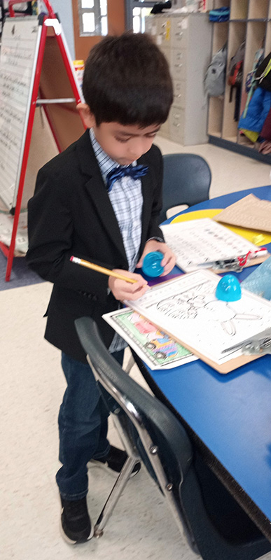A second=grade boy, wearing a plaid shirt and blue bow tie, dark jacket and jeans, holds half of a blue plastic egg as he writes on a worksheet.