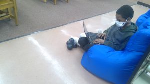 A student sits on a soft blue chair working on a Chromebook.